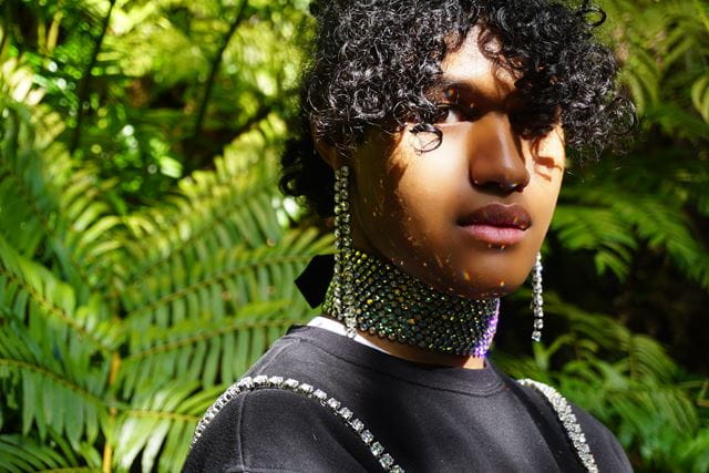 A glamorous headshot of Manu Vaea, a Tongan artist, who stands in front of lush green ferns, wearing sparkly jewellery and a black tshirt